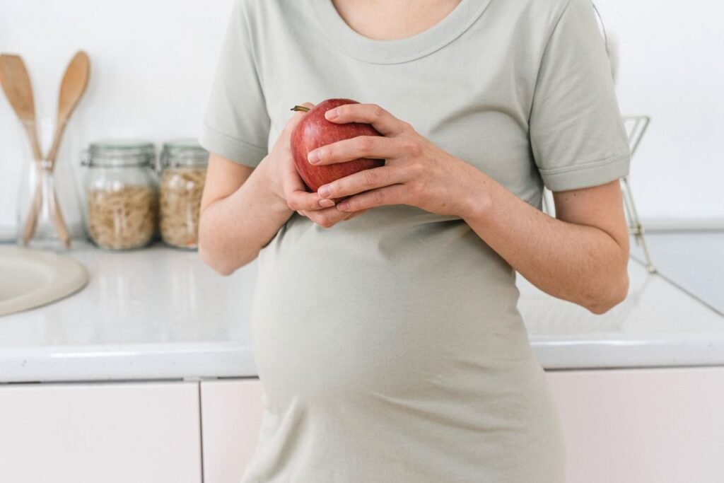 pexels-photo-6991894-6991894 Unrecognizable pregnant female with ripe red apple for healthy diet in hands standing near counter in light kitchen at home